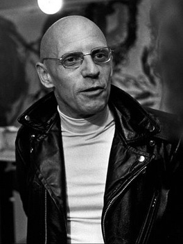 photographie de Michel Foucault, le crâne ras, portant des lunettes, un col roulé blanc et une veste en cuir noir
